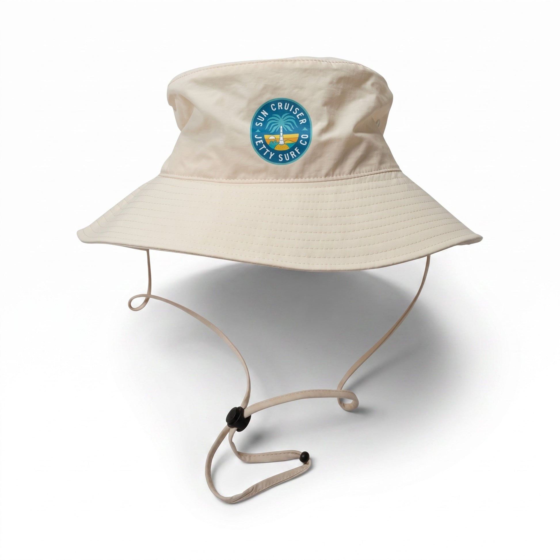 A beige wide-brim bucket hat with an adjustable chin strap, featuring a circular blue and green 'Sun Cruiser x Jetty Surf co' logo  on the front, set against a plain white background.