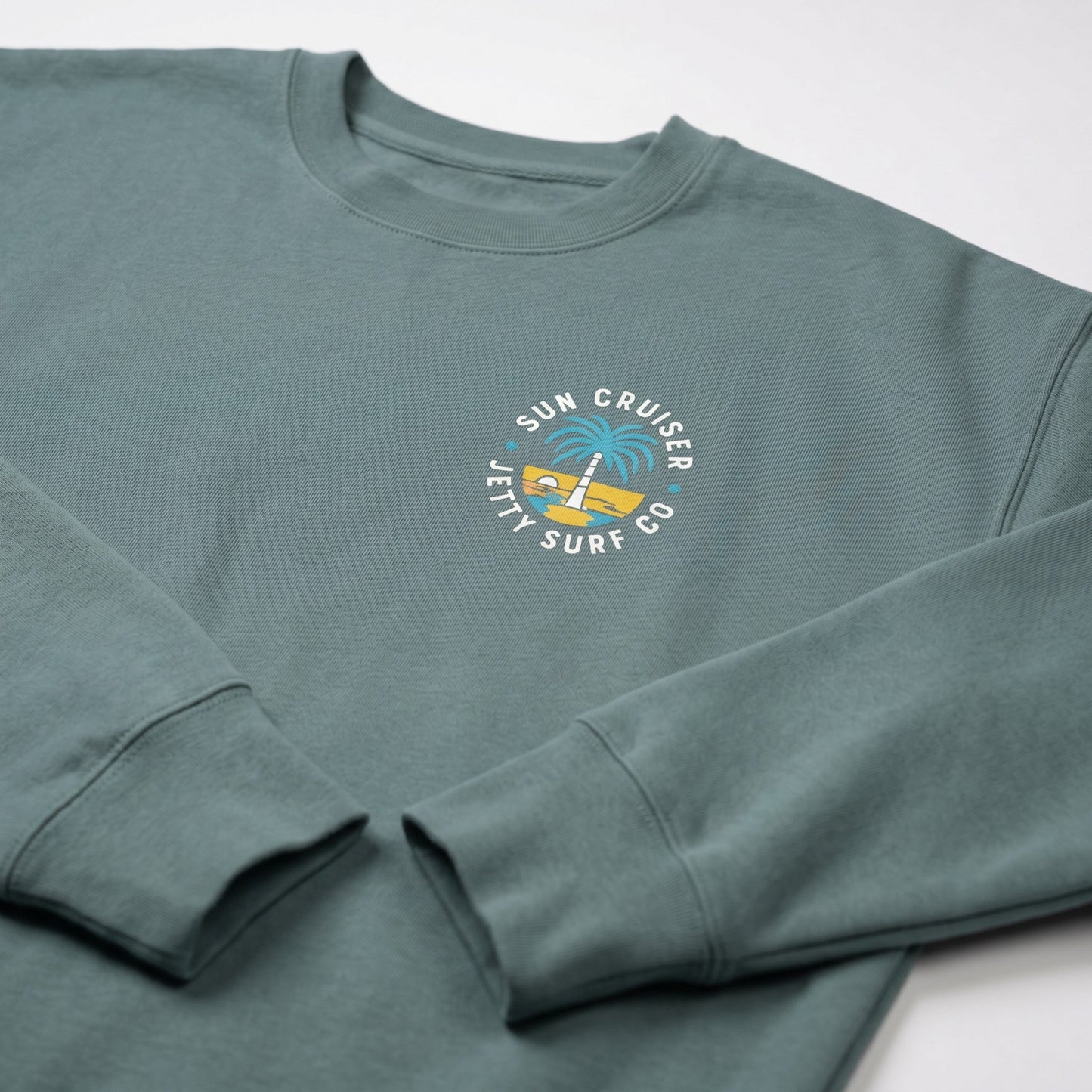 A sage green crewneck sweatshirt with a circular graphic on the left chest reading 'Sun Cruiser Jetty Surf Co' around a yellow and blue palm tree sunset illustration, shown on a light gray background.