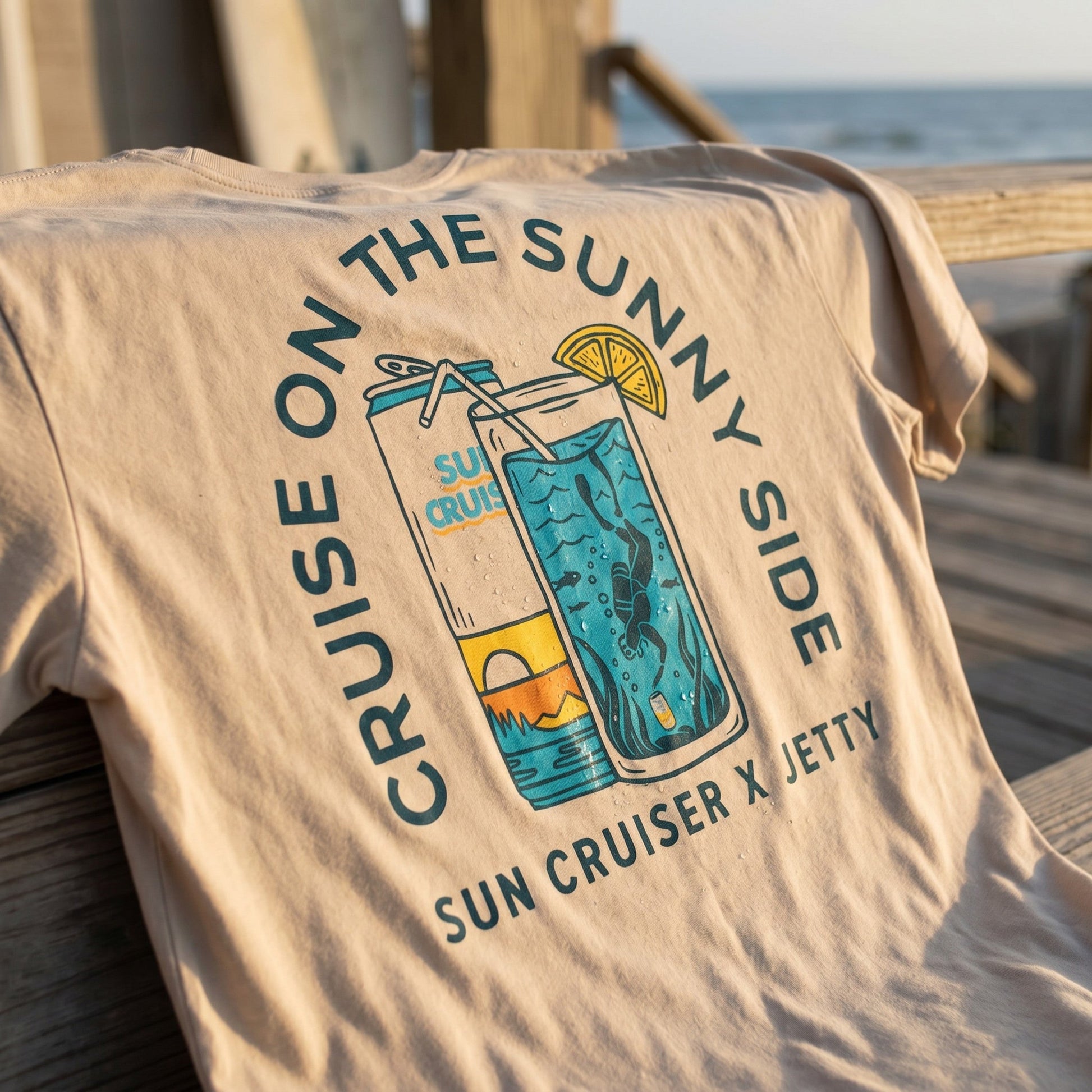 A beige short-sleeve T-shirt draped over a wooden railing at the beach, featuring a large graphic print of a can and a cocktail glass with the text 'CRUISE ON THE SUNNY SIDE' and 'SUN CRUISER X JETTY'.