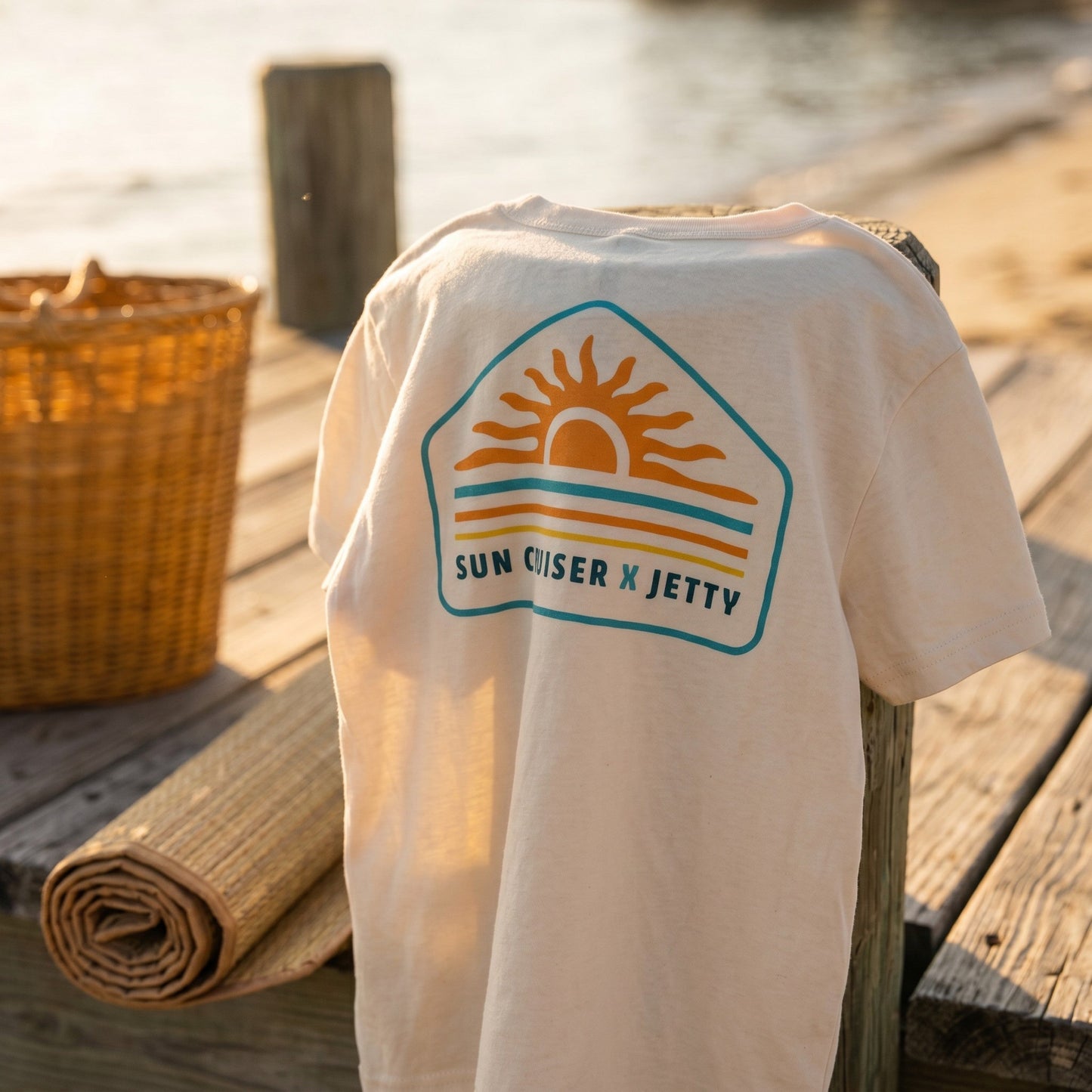 A white t-shirt draped over a wooden post on a dock, featuring a retro-style 'Sun Cruiser x Jetty' graphic in orange, yellow, and teal. The background shows a warm, blurred beach and water scene with a woven basket nearby.