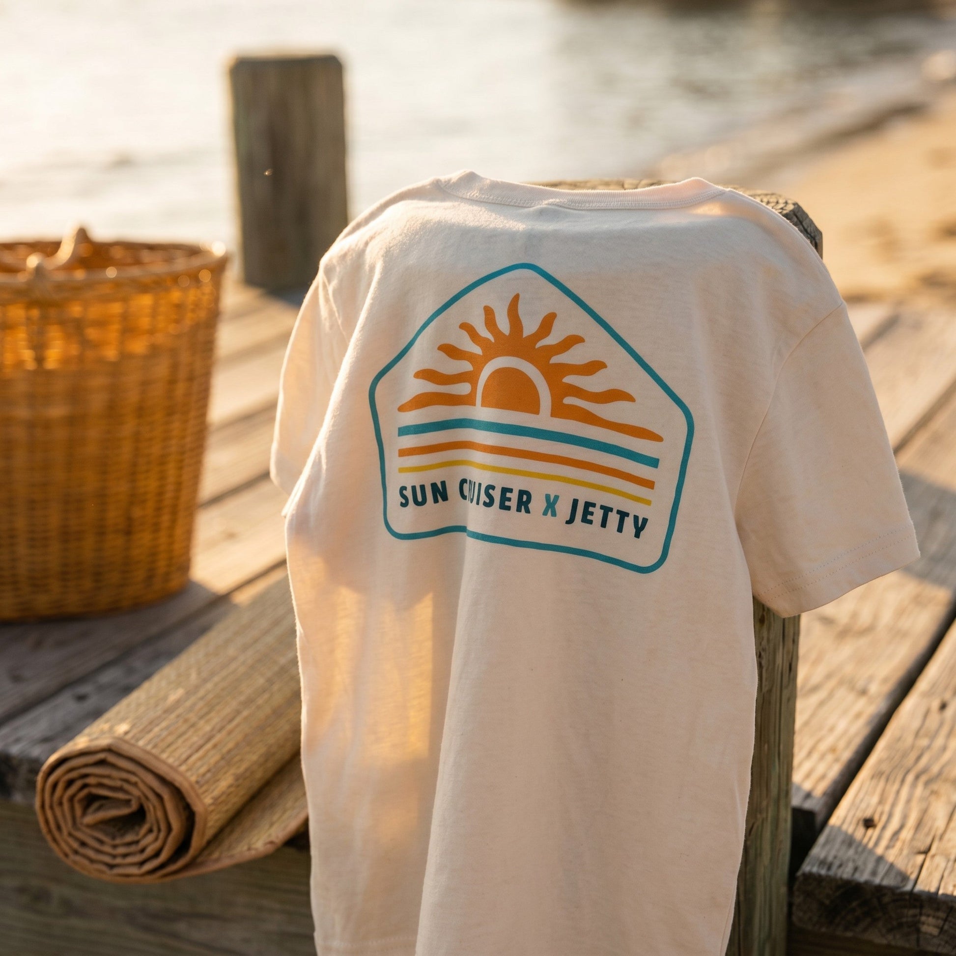 A white t-shirt draped over a wooden post on a dock, featuring a retro-style 'Sun Cruiser x Jetty' graphic in orange, yellow, and teal. The background shows a warm, blurred beach and water scene with a woven basket nearby.