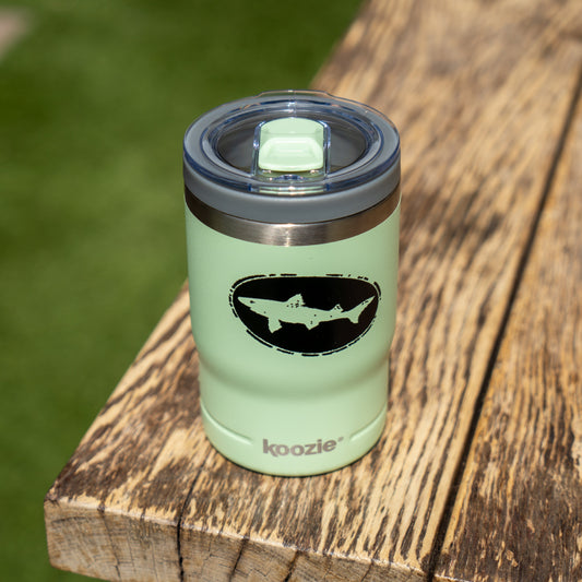 The Sage 12oz Can Insulator by Arrasmith Promotions LLC, featuring a black shark logo, sits on a rustic wooden surface with blurred green grass in the background. Its vacuum-insulated design keeps drinks perfectly chilled.