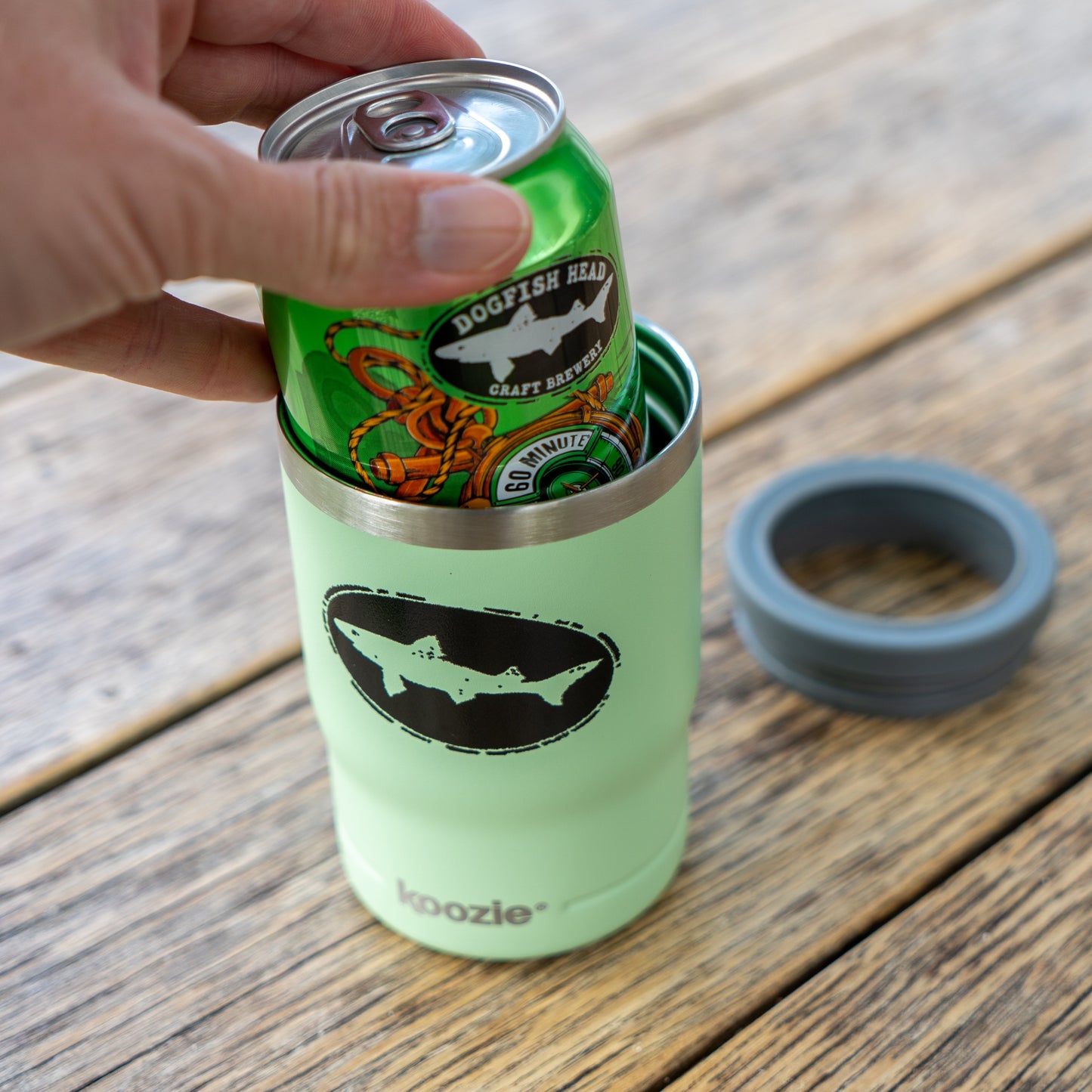A hand places a green Dogfish Head 60 Minute IPA can into an Arrasmith Promotions LLC Sage 12oz Can Insulator on a wooden surface, with the insulator’s removable gray lid beside it.