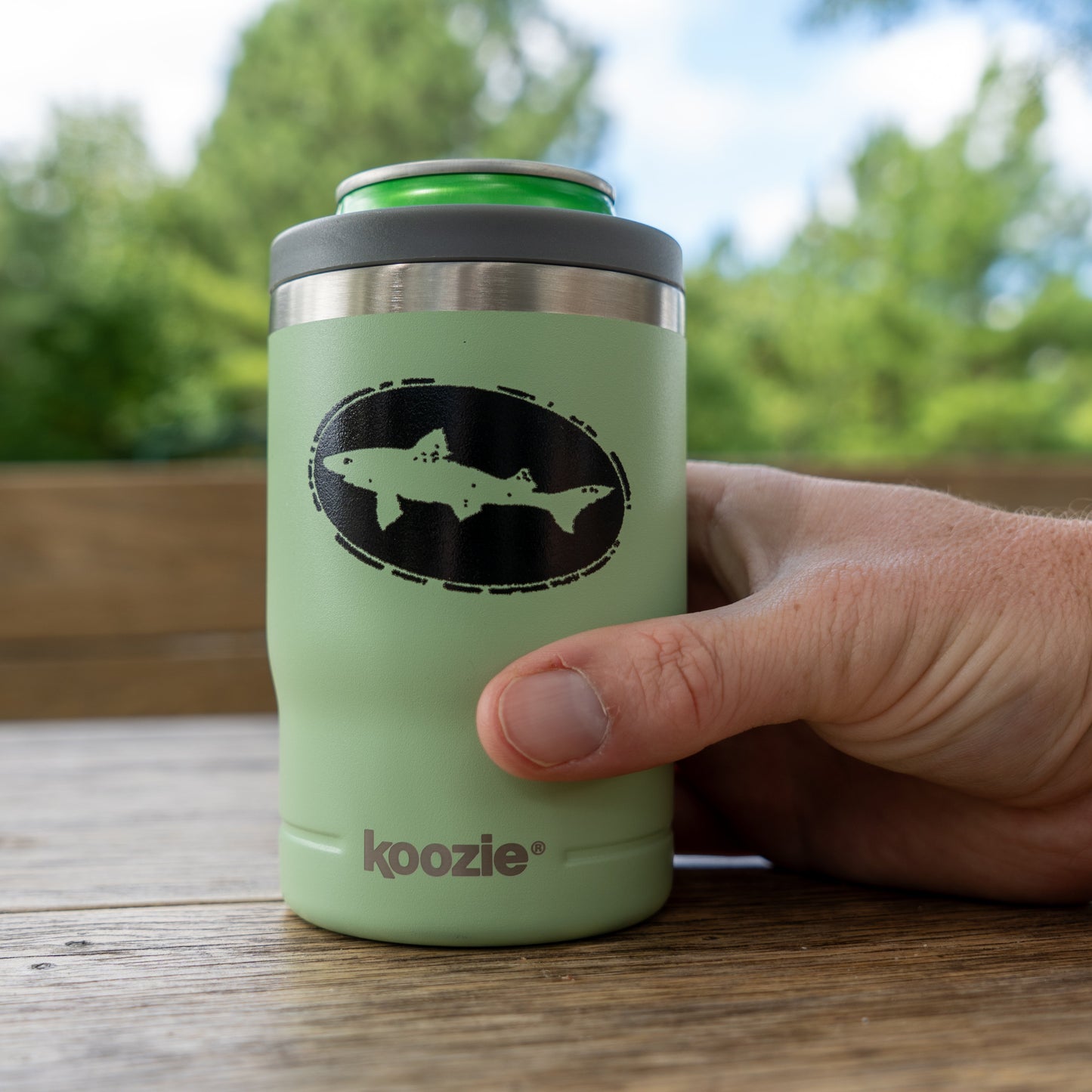 A hand holds the Sage 12oz Can Insulator by Arrasmith Promotions LLC, featuring a green design with a black fish logo and a green can inside. The outdoor setting with trees and wood highlights this stylish vacuum-insulated can cooler.