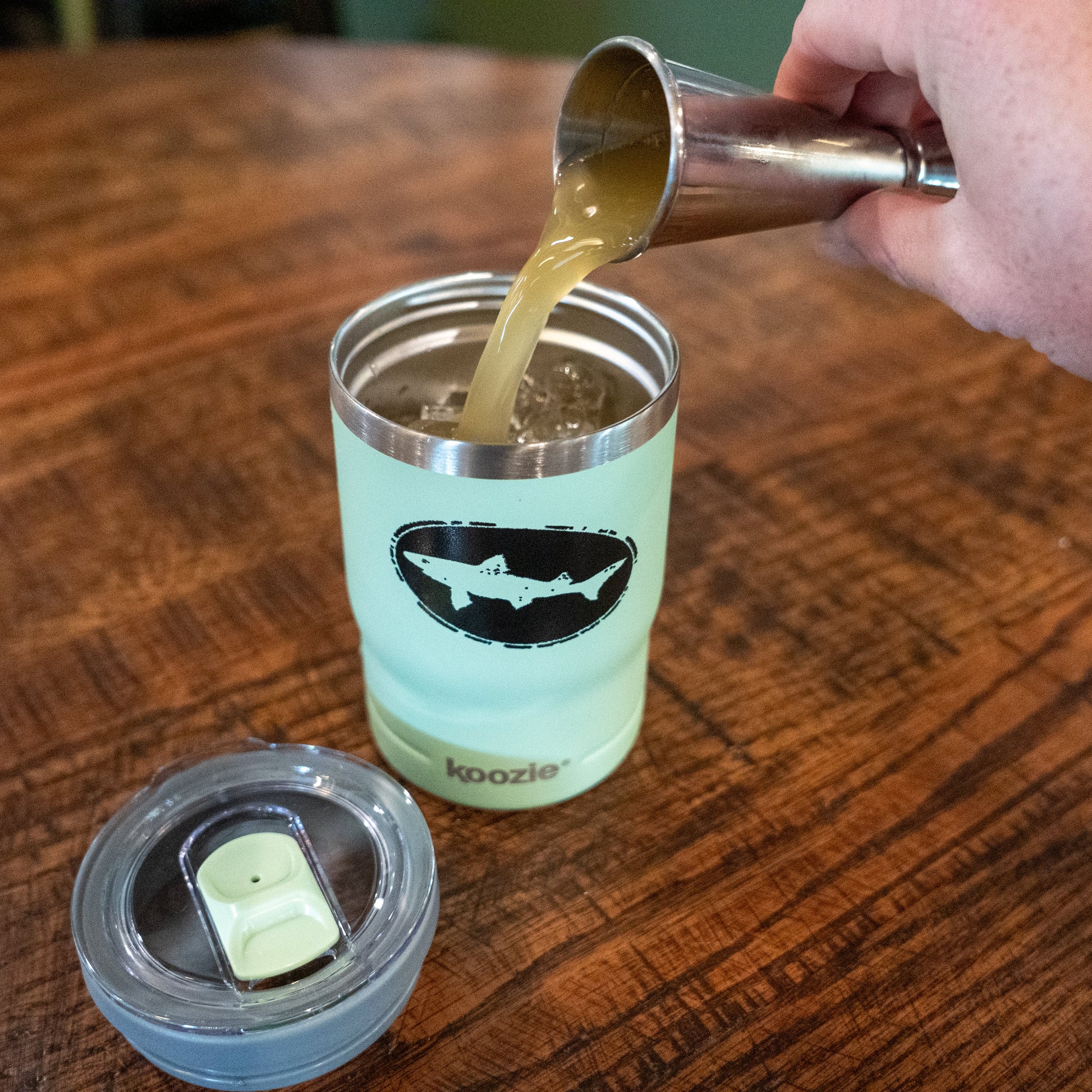 A hand pours a light brown liquid from a metal jigger into a Sage 12oz Can Insulator by Arrasmith Promotions LLC—a light green, vacuum-insulated can cooler with a shark logo—resting on a wooden table. The clear lid sits nearby.