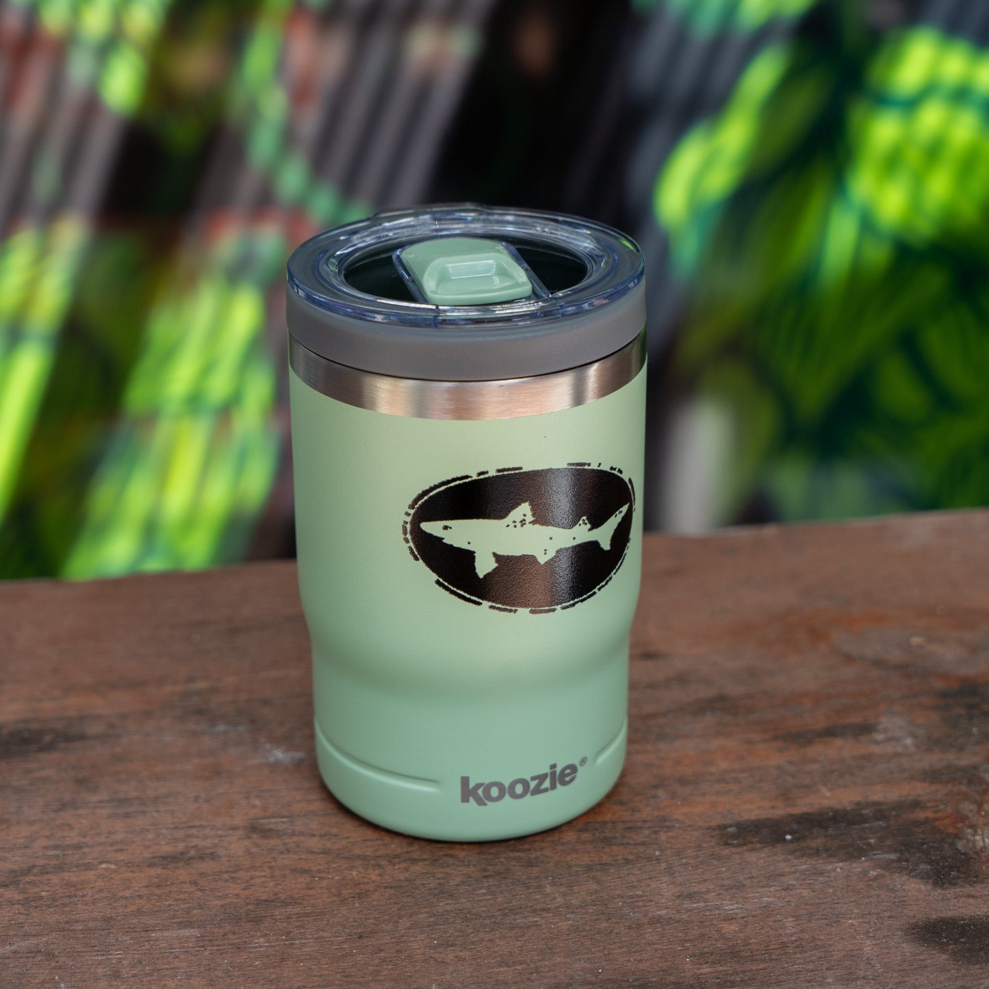 A Sage 12oz Can Insulator from Arrasmith Promotions LLC in light green stainless steel, featuring a black shark logo and "koozie" text, rests on a wooden surface with blurred green and yellow foliage in the background.