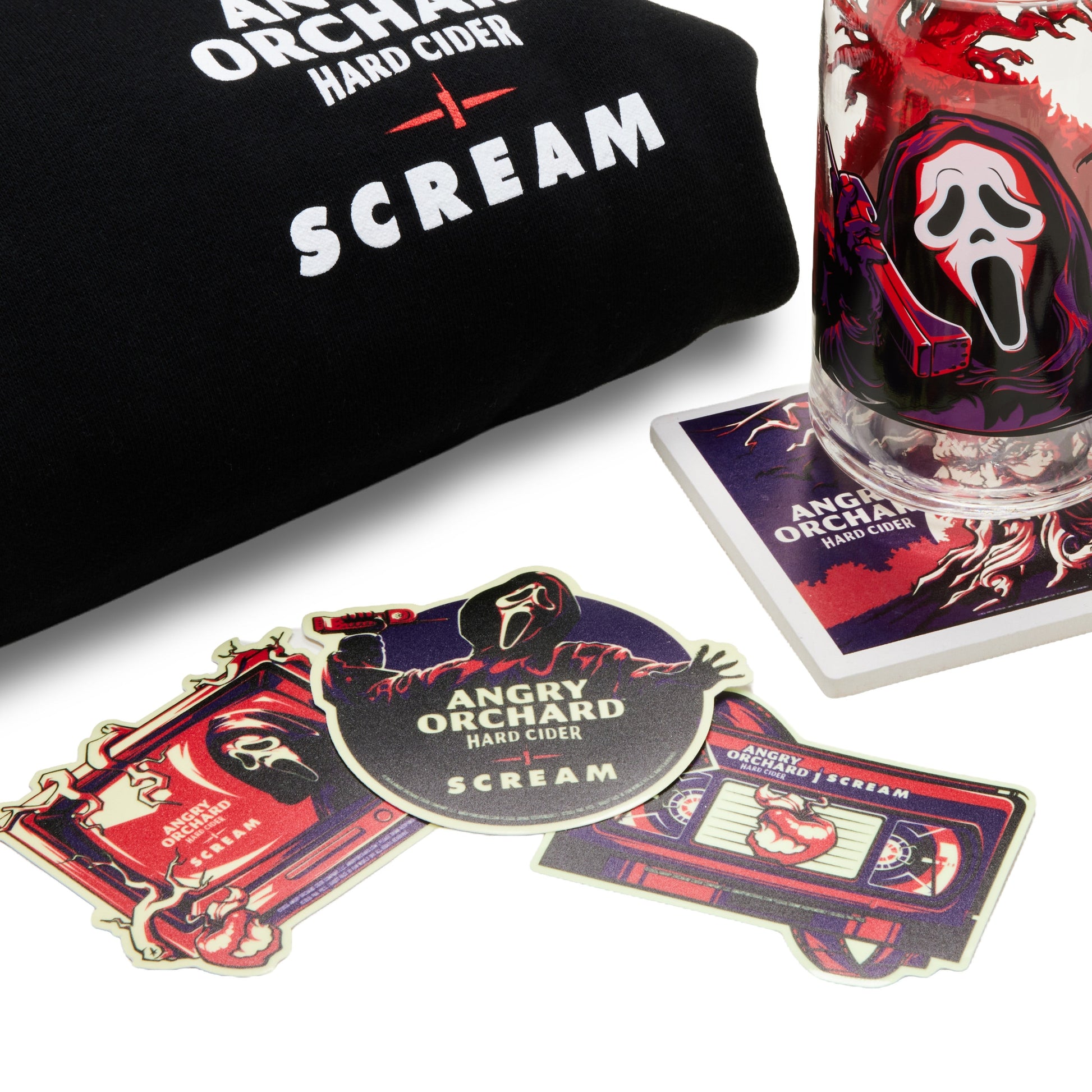 Angry Orchard Hard Cider x Scream collaboration merchandise on a white background, including a black t-shirt with white text, a glass mug featuring the Ghostface graphic, a coaster, and three die-cut stickers.
