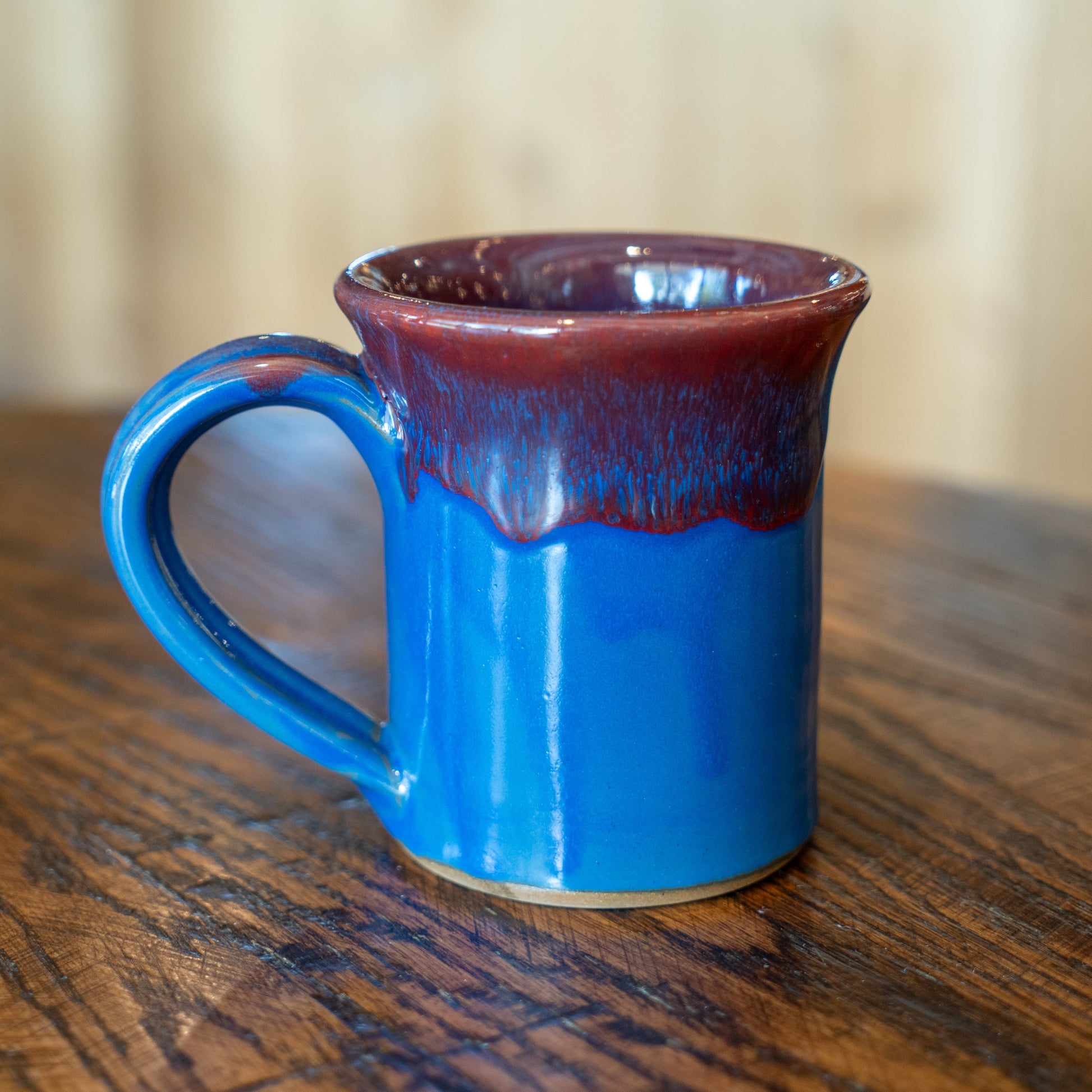 The Deneen Pottery Sky Blue Core Flare Ceramic Mug, featuring a glossy maroon rim and a rounded handle, sits off-center on a wooden table with blurred wood paneling in the background.