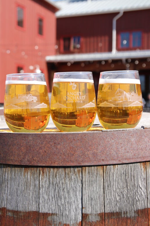 On one side of this glass is an apple and under it the writing "Angry Orchard Hard Cider". Under the writing is the beginning of a silhouette of the Angry Orchard, which continues to the next side. The other side of the glass as a full white silhouette of the Angry Orchard. 