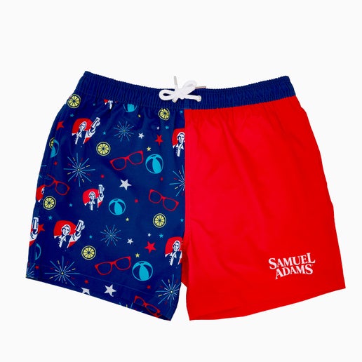 Split design swim trunks. One half is dark blue with red, white, and blue fireworks, stars, beachballs, and Samuel Adams logo on it. The other half is solid red with a white samuel adams logo on bottom of the trunk leg