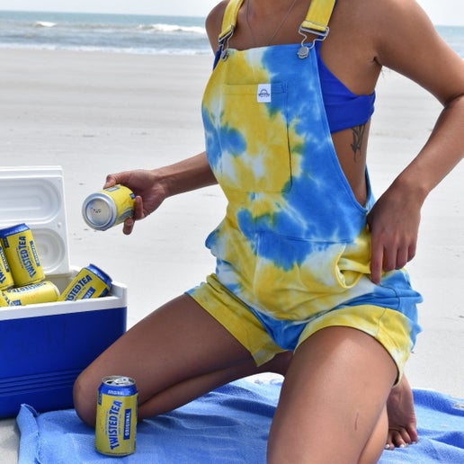 Person wearing blue and yellow tie‑dye short overalls while sitting on a beach towel near the ocean, holding a Twisted Tea Original can beside an open cooler filled with ice and Twisted Tea cans.