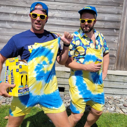 Two people wearing blue and yellow tie‑dye short overalls outdoors, holding Twisted Tea cans and packaging, standing in front of a wooden fence on grass.