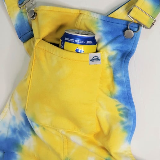 Close‑up of blue and yellow tie‑dye short overalls showing the front bib pocket holding a Twisted Tea hard iced tea can, with adjustable shoulder straps and metal hardware visible.