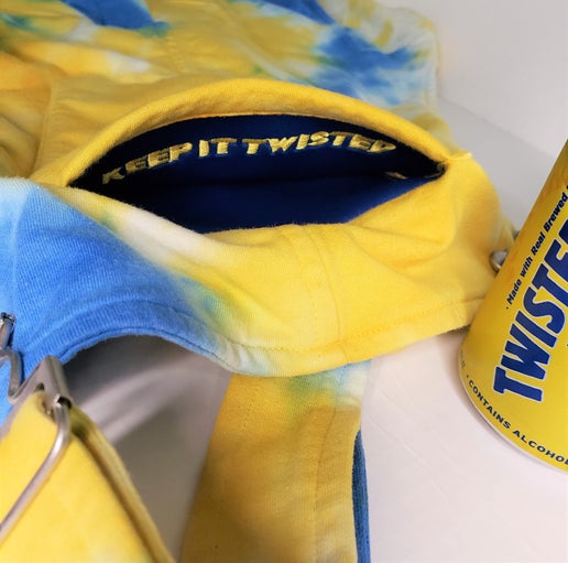 Close‑up of the interior pocket of blue and yellow tie‑dye overalls showing navy lining with embroidered “Keep It Twisted” text, alongside a Twisted Tea can