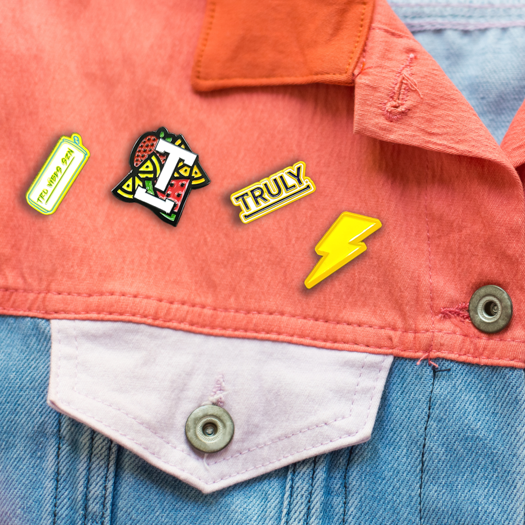 A close-up view of a denim jacket's peach-colored top and light blue denim bottom, adorned with four Truly-themed enamel pins. The pins include a yellow "TRU VIBES SZN" can, a stylized "T" with a fruit pattern, a yellow "TRULY" pin, and a yellow lightning bolt.