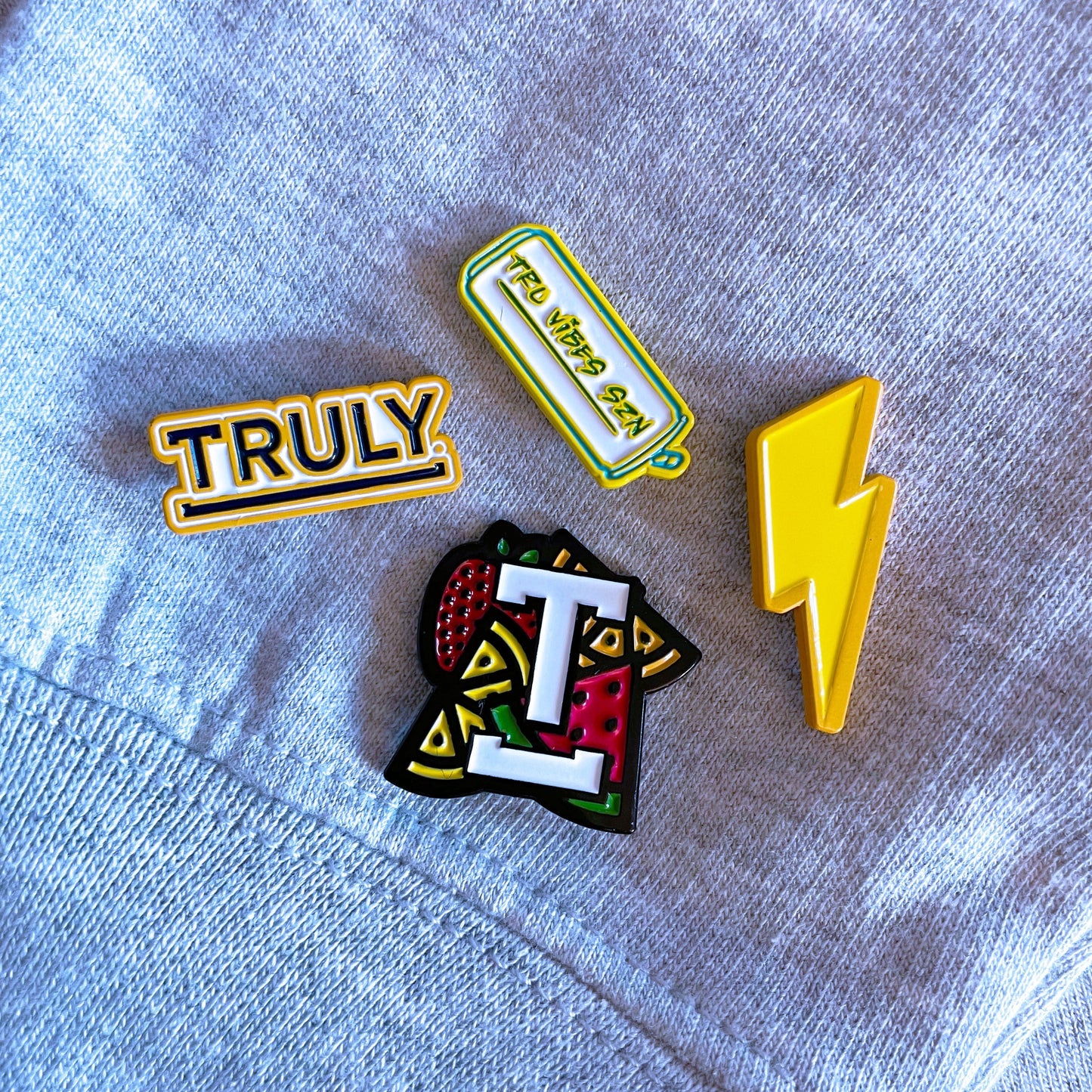 A close-up view of a blue piece of fabric adorned with four Truly-themed enamel pins. The pins include a yellow rectangular pin with "TRULY", a yellow can-shaped pin with "TRU VIBES SZN", a yellow lightning bolt pin, and a stylized "T" pin with a fruit pattern.