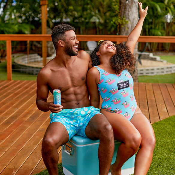 A man with a beard and a shirtless, torso sits on a blue and white cooler, holding a Truly Hard Seltzer can. To his right, a woman with curly hair wears a light blue swimsuit with a flamingo and "TRULY" pattern, raising her right arm in the air. They are outdoors on a patio with grass and a tree in the background.