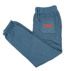 A pair of dark teal sweatpants is shown. The pants feature an elasticized waistband with a drawstring for adjustment. Side pockets are visible on each hip, and a patch pocket is located on the rear right side. In red on the center of the back pocket is the word "TRULY" in a stylized, font. The legs taper slightly towards the ribbed cuffs at the ankles. The fabric appears to be a soft, comfortable knit.