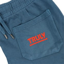 A close-up shot focuses on the rear right pocket of a pair of dark blue sweatpants. The patch pocket is clearly visible, and in bright red, capital letters on the center of the pocket is the word "TRULY". A horizontal red line underlines the word. To the left of the pocket, part of the elasticized waistband and the drawstring with silver aglets are visible. 
