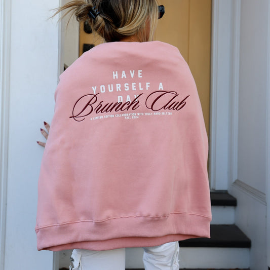Back of a person wearing a pink sweatshirt with the words 'HAVE YOURSELF A DAY Brunch Club' in white and brown script. Below, in smaller text, it reads 'A LIMITED EDITION COLLABORATION WITH TRULY HARD SELTZER FALL 2024'.