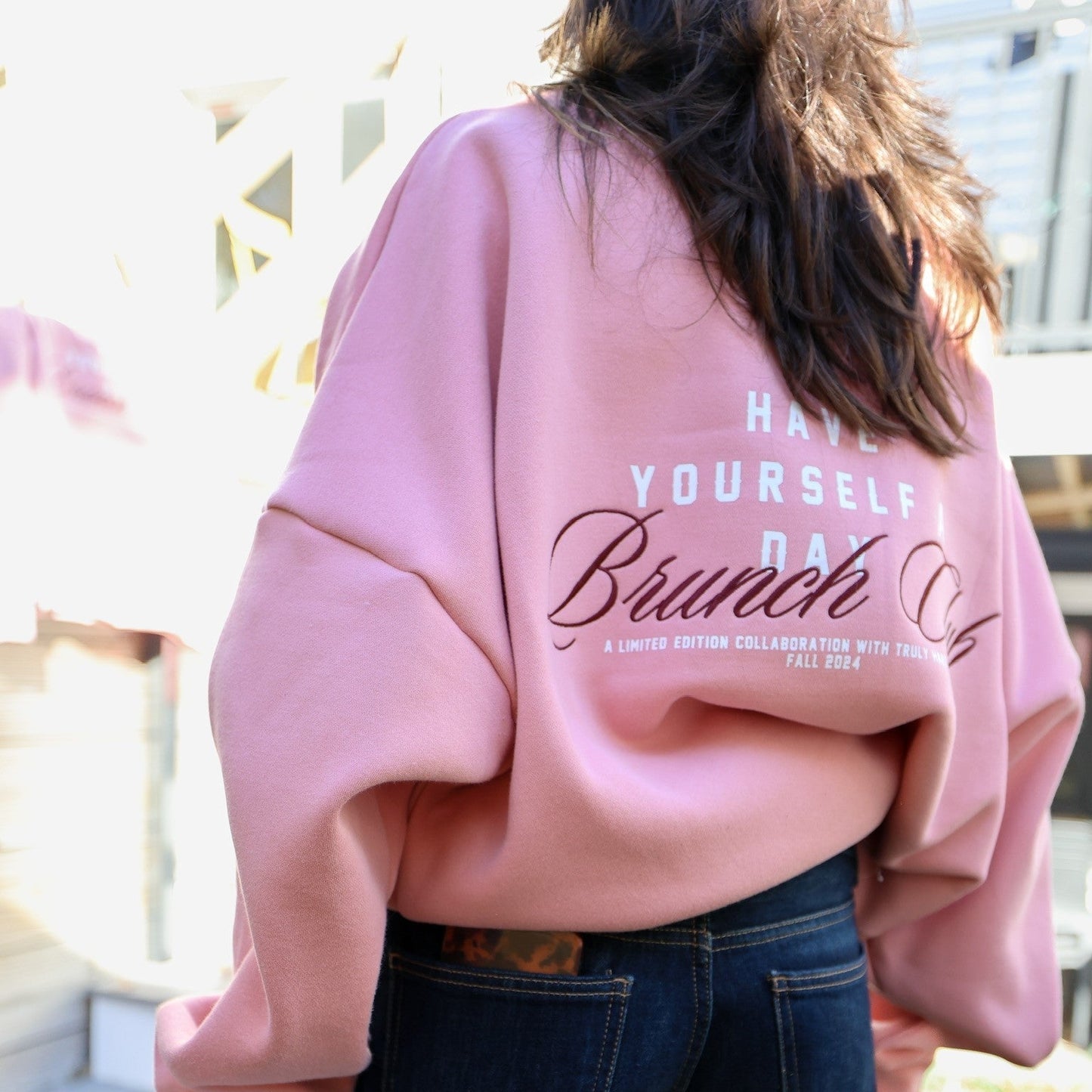 Back of a person wearing an oversized pink sweatshirt with 'HAVE YOURSELF A DAY Brunch Club' printed in white and brown. Below, in smaller text, it reads 'A LIMITED EDITION COLLABORATION WITH TRULY HARD SELTZER FALL 2024'. The person is also wearing dark jeans, and there's a light-colored railing or banister in the blurred background.