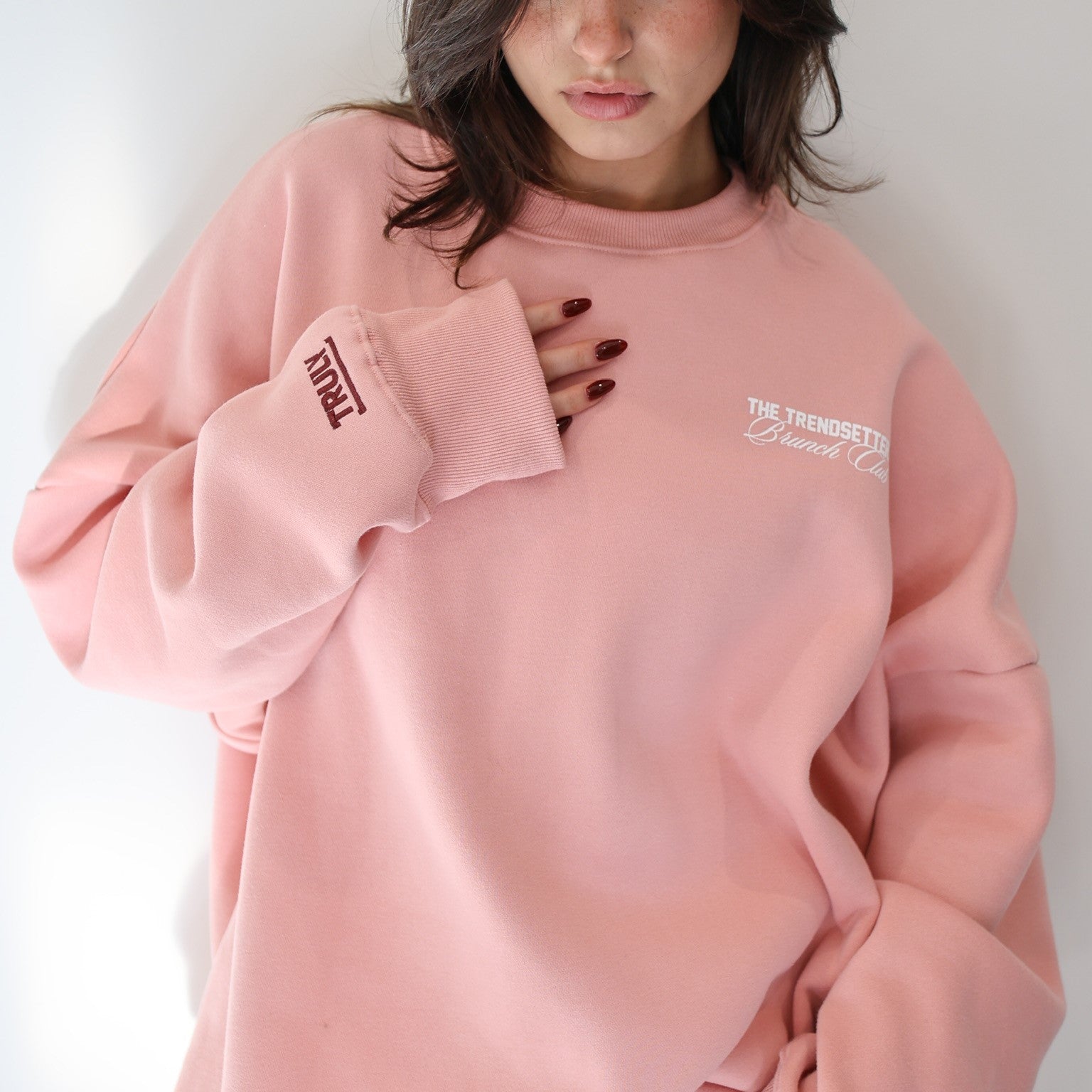 Front view of a person wearing a pink crewneck sweatshirt. The right cuff of the sweatshirt has 'TRULY' printed vertically in dark red. On the left chest area, 'THE TRENDSETTER Brunch Club' is printed in white script. The person's left hand is resting near their chest, and they have dark red nail polish.