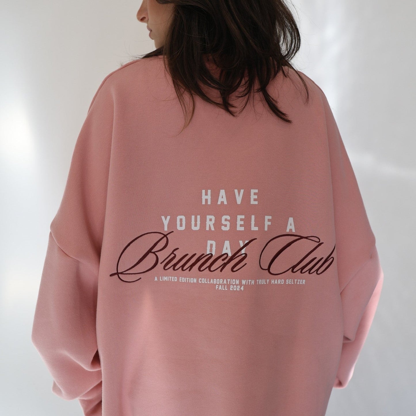 Back of a person wearing a pink crewneck sweatshirt. The back of the sweatshirt features a large design with 'HAVE YOURSELF A DAY Brunch Club' in white and brown script. Below this, in smaller text, it reads 'A LIMITED EDITION COLLABORATION WITH TRULY HARD SELTZER FALL 2024'.