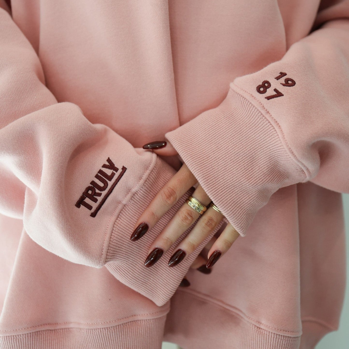 Close-up of a person's hands and the cuffs of a pink sweatshirt. The left cuff has 'TRULY' printed vertically in dark red. The right cuff has the numbers '19' and '87' stacked vertically in dark red. The person has dark red nail polish and is wearing two rings on their left hand.