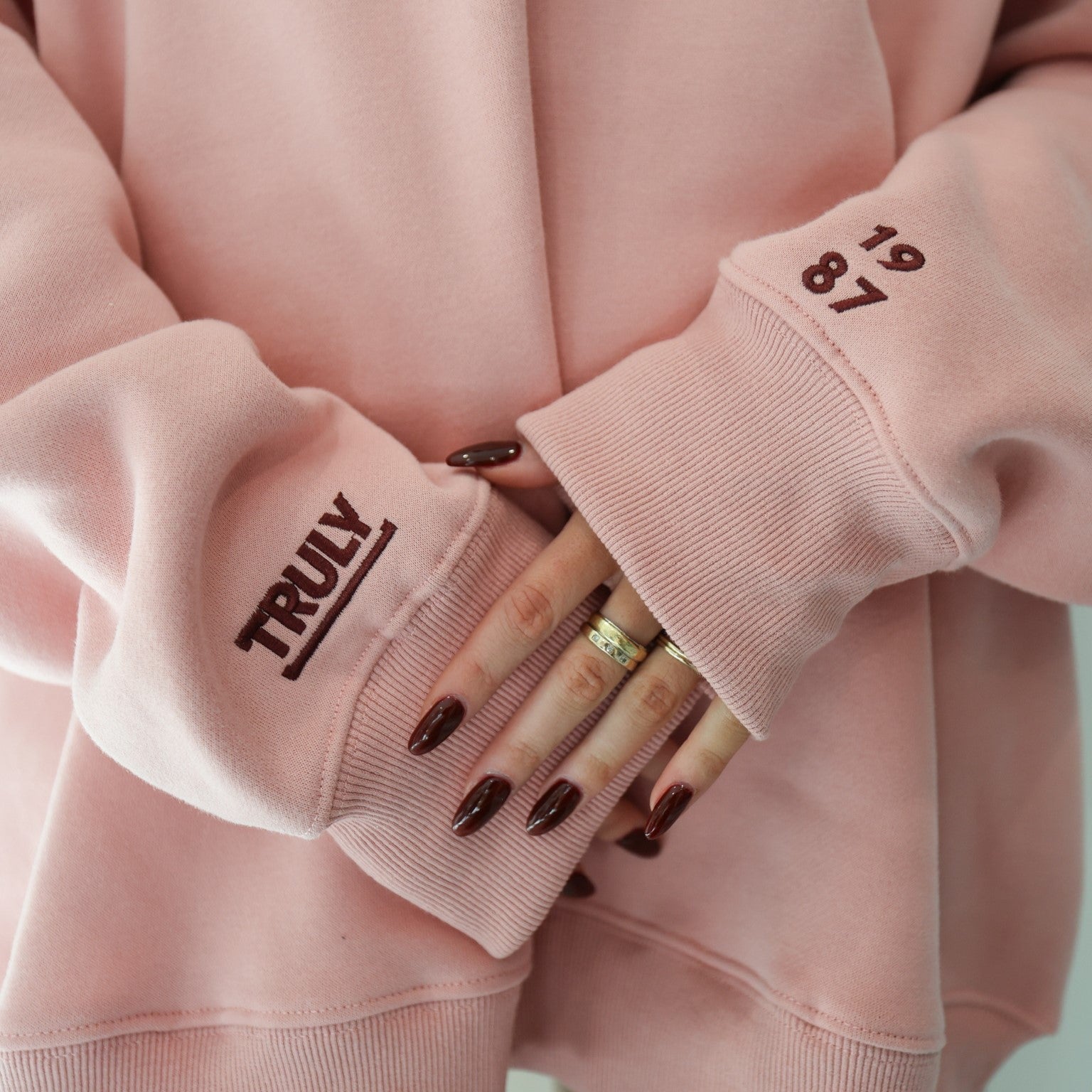 Close-up of a person's hands and the cuffs of a pink sweatshirt. The left cuff has 'TRULY' printed vertically in dark red. The right cuff has the numbers '19' and '87' stacked vertically in dark red. The person has dark red nail polish and is wearing two rings on their left hand.