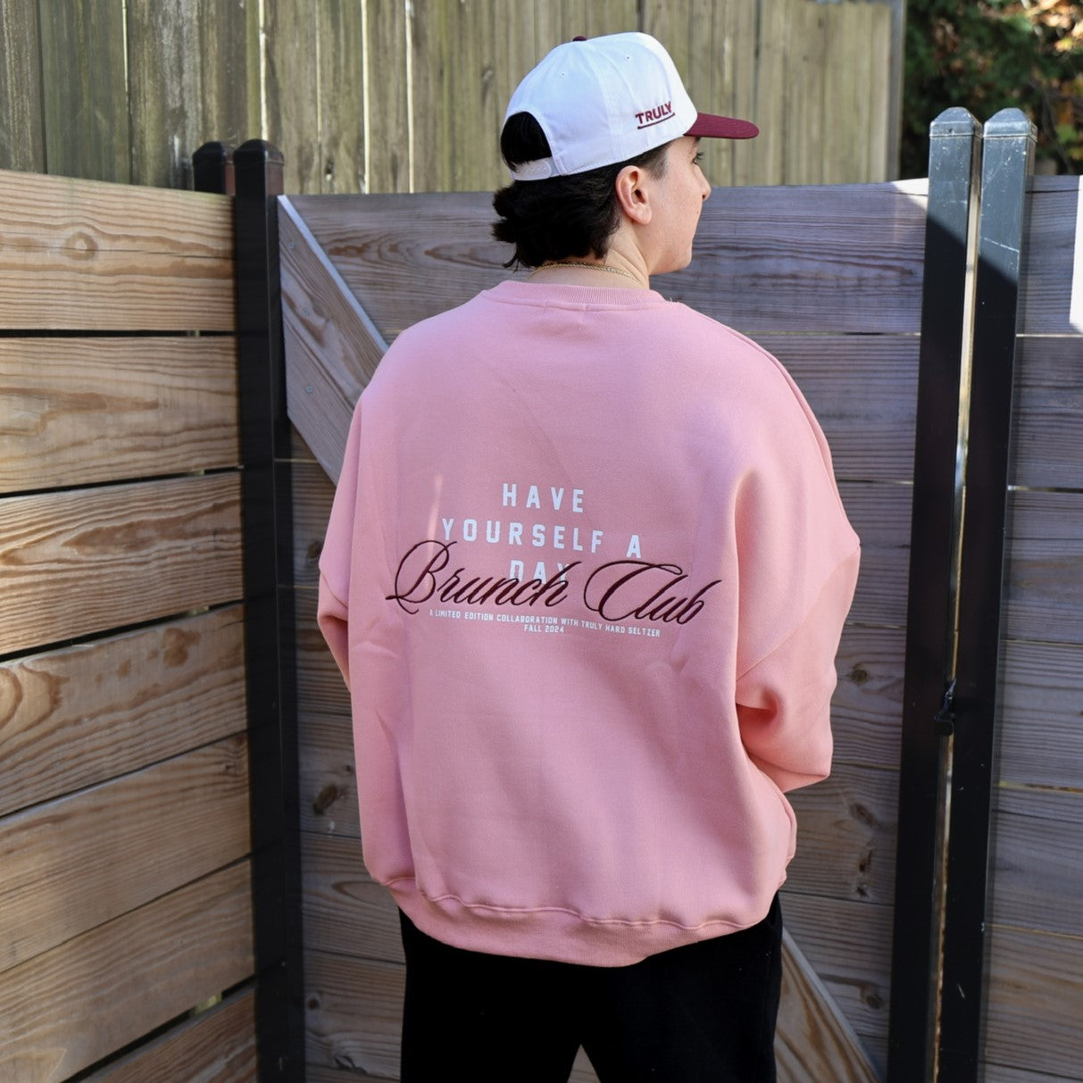 Back of a person standing outdoors, facing slightly right, wearing an oversized pink sweatshirt and a white and maroon baseball cap. The back of the sweatshirt has 'HAVE YOURSELF A DAY Brunch Club' in white and brown text, with smaller text below reading 'A LIMITED EDITION COLLABORATION WITH TRULY HARD SELTZER FALL 2024'. The brim of the cap shows 'TRULY'. The person is standing in front of a rustic wooden fence.