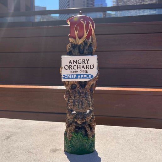 This tall tap handle is the Angry Orchard tree trunk with a face on it. It has green grass at the bottom and the iconic Angry Orchard red bitten apple on the top. Below the apple is a white board with writing "Angry Orchard Hard Cider" "Crisp Apple" on it.