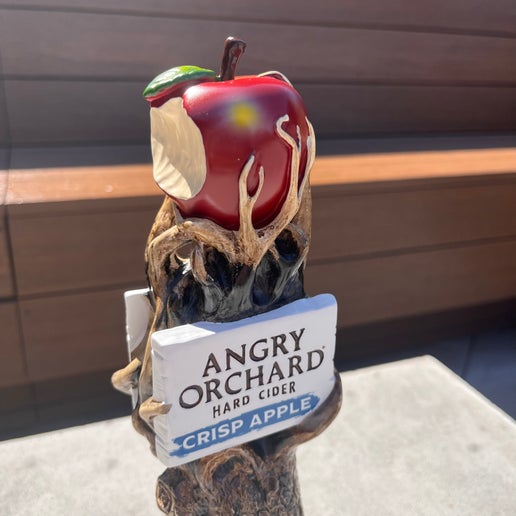 This tall tap handle is the Angry Orchard tree trunk with a face on it. It has green grass at the bottom and the iconic Angry Orchard red bitten apple on the top. Below the apple is a white board with writing "Angry Orchard Hard Cider" "Crisp Apple" on it.