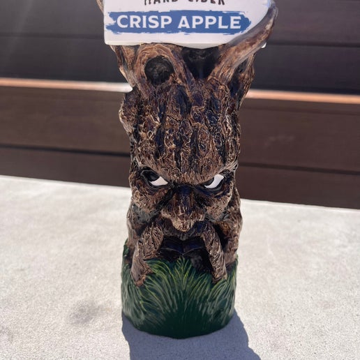 This tall tap handle is the Angry Orchard tree trunk with a face on it. It has green grass at the bottom and the iconic Angry Orchard red bitten apple on the top. Below the apple is a white board with writing "Angry Orchard Hard Cider" "Crisp Apple" on it.