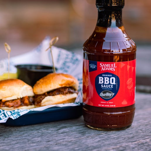 The Sam Adams x CharBoy's BBQ Sauce bottle shown in front of two sandwiches