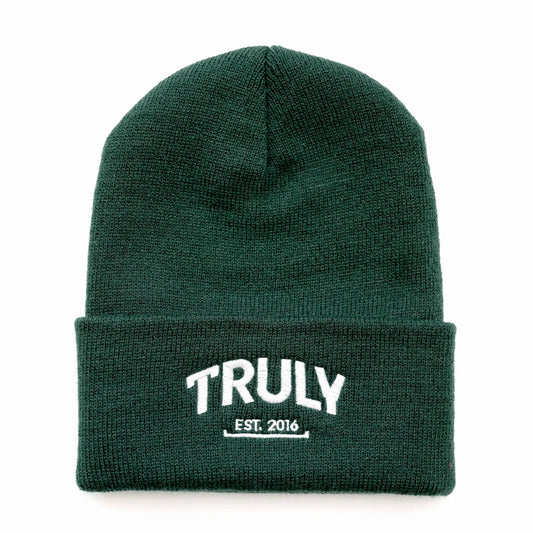 A dark green knit beanie with a folded cuff. "TRULY" is embroidered in white letters with "EST. 2016" underneath, also in white.