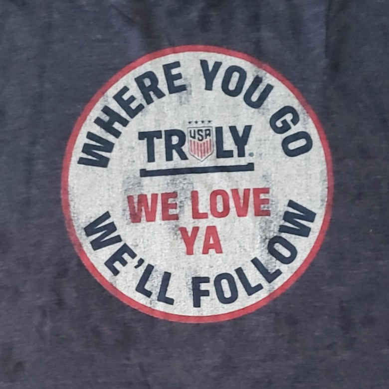 A close-up of a distressed circular logo on a t-shirt. The logo has a red outline and a faded, white background with a worn appearance. Around the top half of the circle, in dark gray capital letters, it reads "WHERE YOU GO". Below this, centered horizontally, is "TRULY" in dark gray capital letters with a small "USA" shield logo and a thin dark gray line underneath. Below the "TRULY" text, in red capital letters, it says "WE LOVE YA". The bottom of the circle, in gray, it reads "WE'LL FOLLOW".