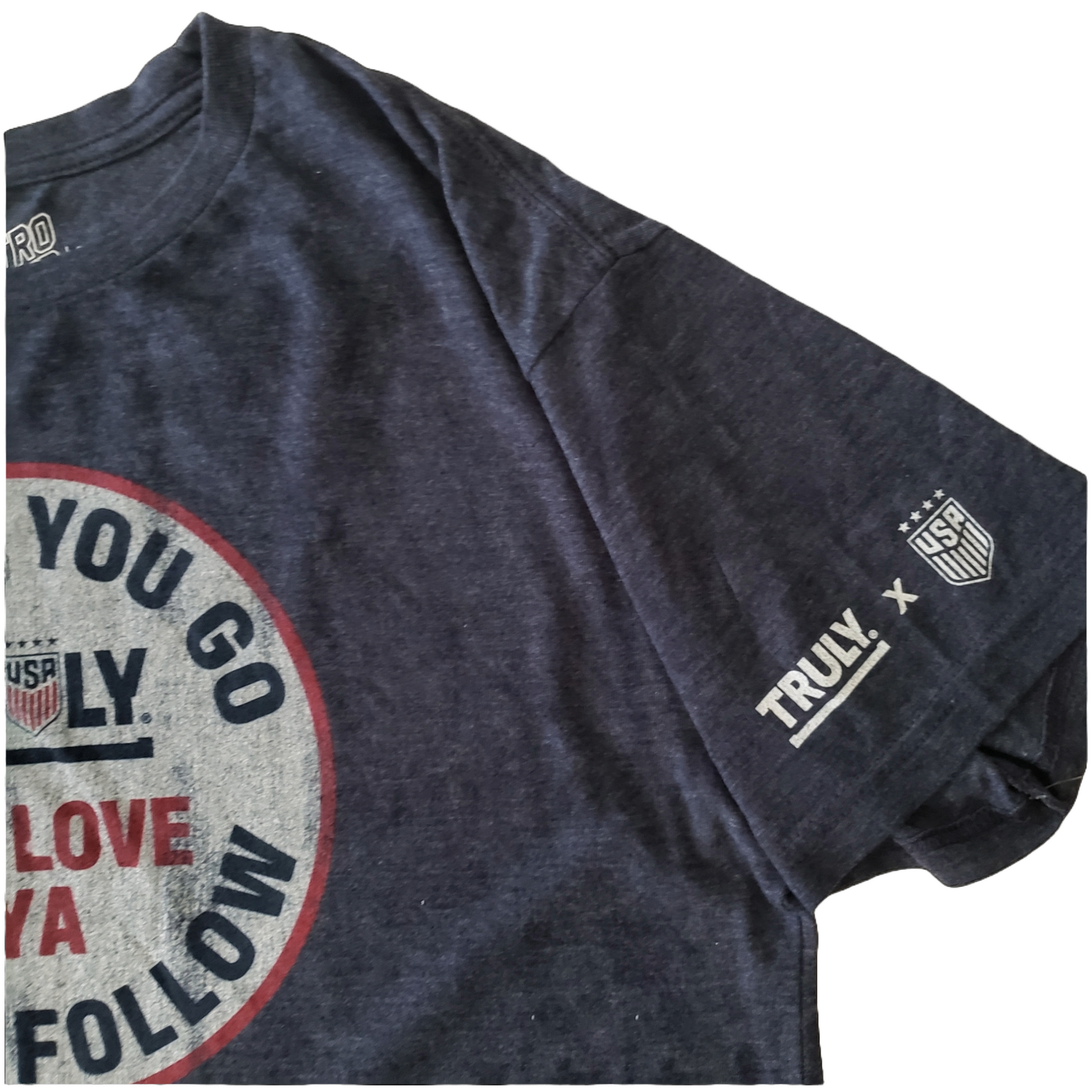 
A dark gray t-shirt with a portion of a distressed circular logo visible on the left side of the chest. The logo has a red border and a faded white background with the words "WHERE YOU GO WE'LL FOLLOW" in dark gray around the edge, and "TRULY" with a "USA" shield logo, and "WE LOVE YA" in red, in the center. On the left sleeve, near the cuff, there's a white print that reads "TRULY X" followed by a "USA" shield logo with four stars above it.