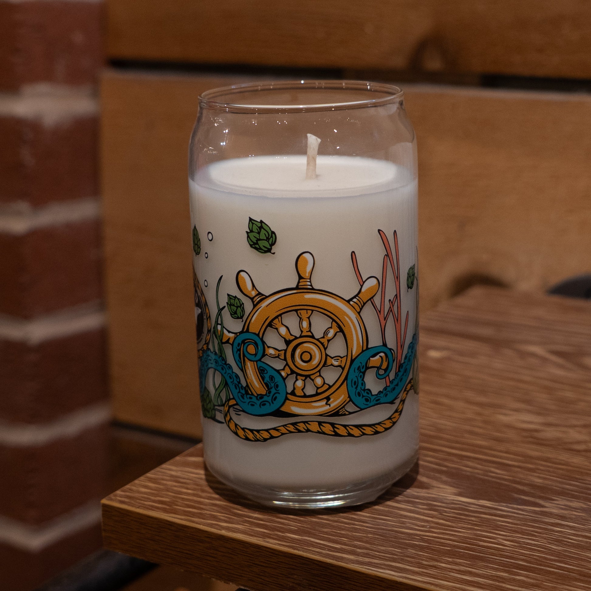 The Under the Sea Can Glass Candle by Unwined Candles, LLC features ocean scents of coconut, mango, and pineapple in a white glass jar with a ship’s wheel, blue waves, green hops, and seaweed on wood against a brick and wood background.