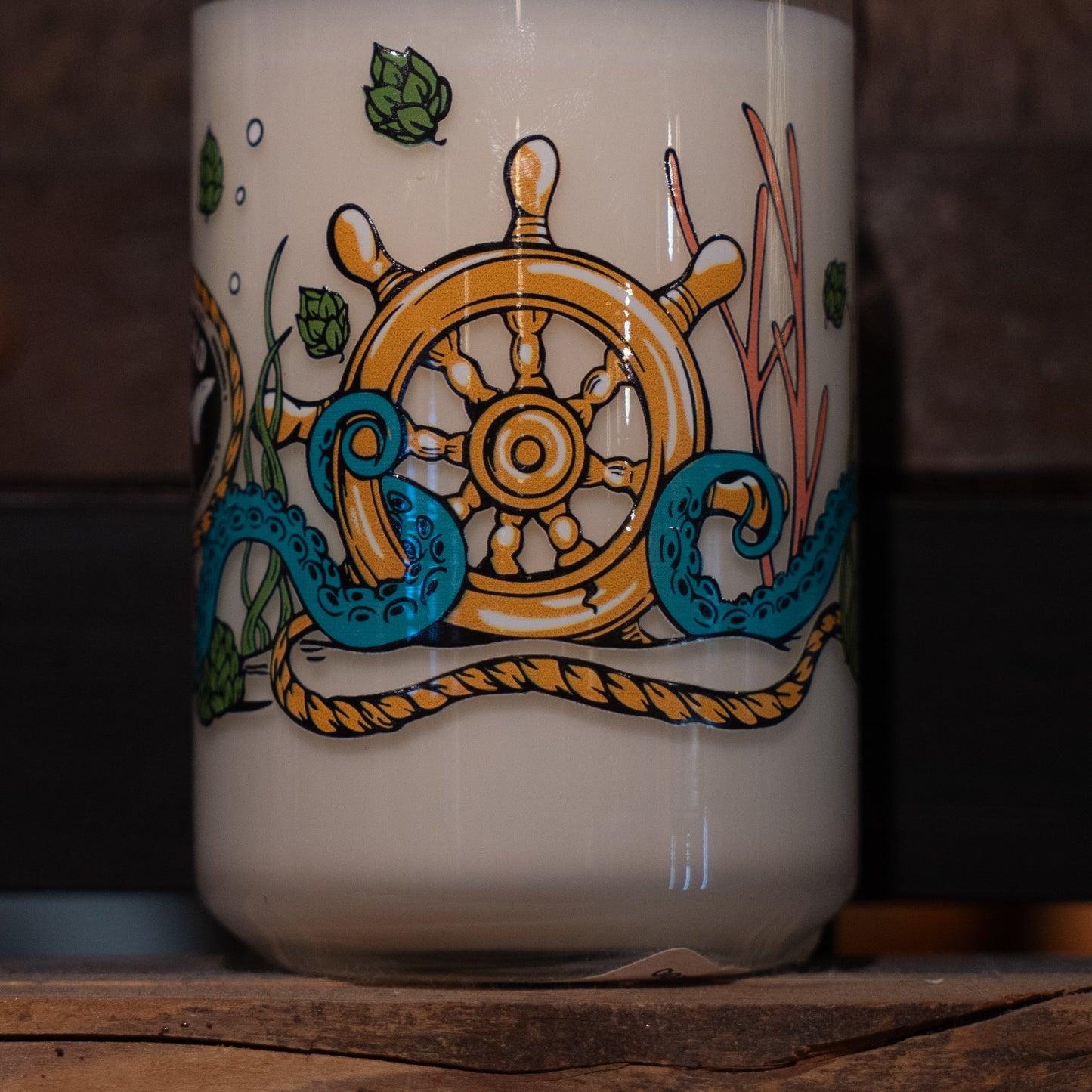 A close-up of the Under the Sea Can Glass Candle by Unwined Candles, LLC, featuring a yellow ship’s wheel, green hops, seaweed, and a blue octopus tentacle—perfect for ocean scent candle fans.