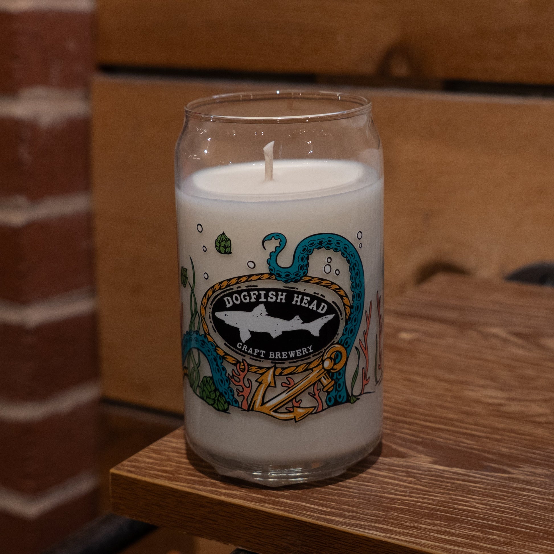 An Under the Sea Can Glass Candle by Unwined Candles, LLC, featuring a teal octopus and underwater scene design, sits on a wooden surface with a wooden and brick background.