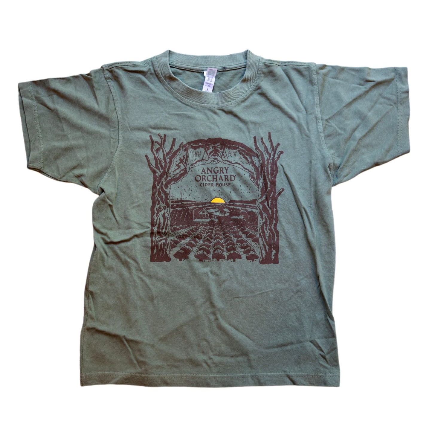 Unisex Green Angry Orchard Treehouse Short Sleeve Shirt. Centered on the front of the shirt is a dark brown hand drawn like image of an orchard with a small yellow sun setting on the horizon. Bordering the sides of this image are two trees that form an arch over the top border imagery. In the center of this imagery reads "Angry Orchard, Cider House" in the classic angry orchard font.