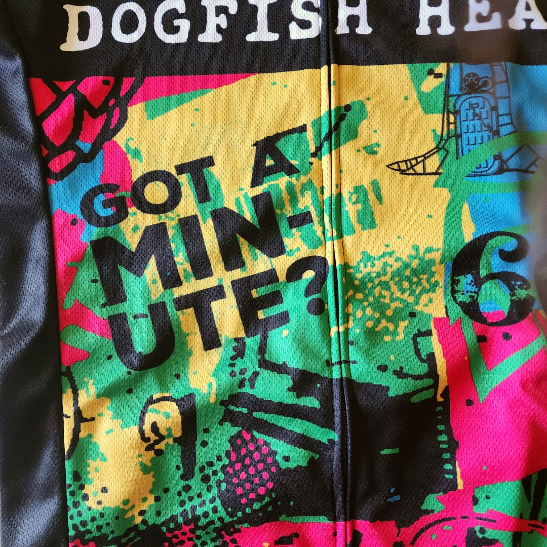 Close-up of Dogfish Head cycling jersey front showing a bright mixed-color design with green, yellow, pink, and blue paint-splatter graphics, bold black text reading ‘Got a Minute?’ across the chest, and a vertical zipper running down the center of the breathable fabric