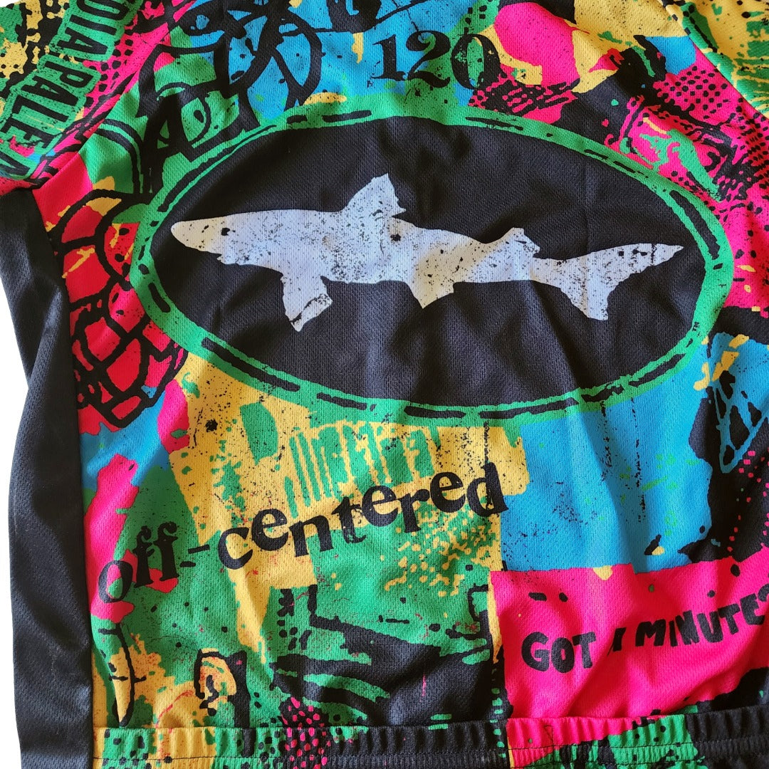 Detailed view of the back of a Dogfish Head bike jersey featuring a large dogfish head logo centered inside a black oval, surrounded by vibrant graffiti-style colors in blue, green, yellow, and pink, with layered abstract illustrations and textured performance material