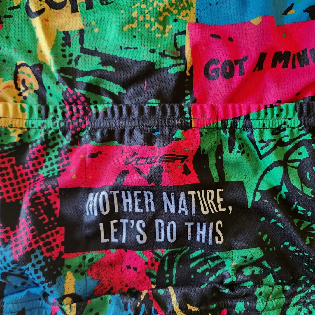 Close-up of the lower back of a colorful Dogfish Head cycling jersey showing stitched rear pockets, multicolor abstract print with black overlays, and white text reading ‘Mother Nature, Let’s Do This’ printed across a black panel on moisture-wicking fabric