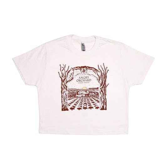 Angry Orchard Cider House design in dark red print on a white bone short sleeve T-shirt. The design is a hand drawn like image of the cider house sitting in the orchard, with a small yellow sun setting on the horizon. Bordering the sides of this image are two trees that form an arch over the top border imagery. In the center of this imagery reads "Angry Orchard, Cider House" in the classic angry orchard font.