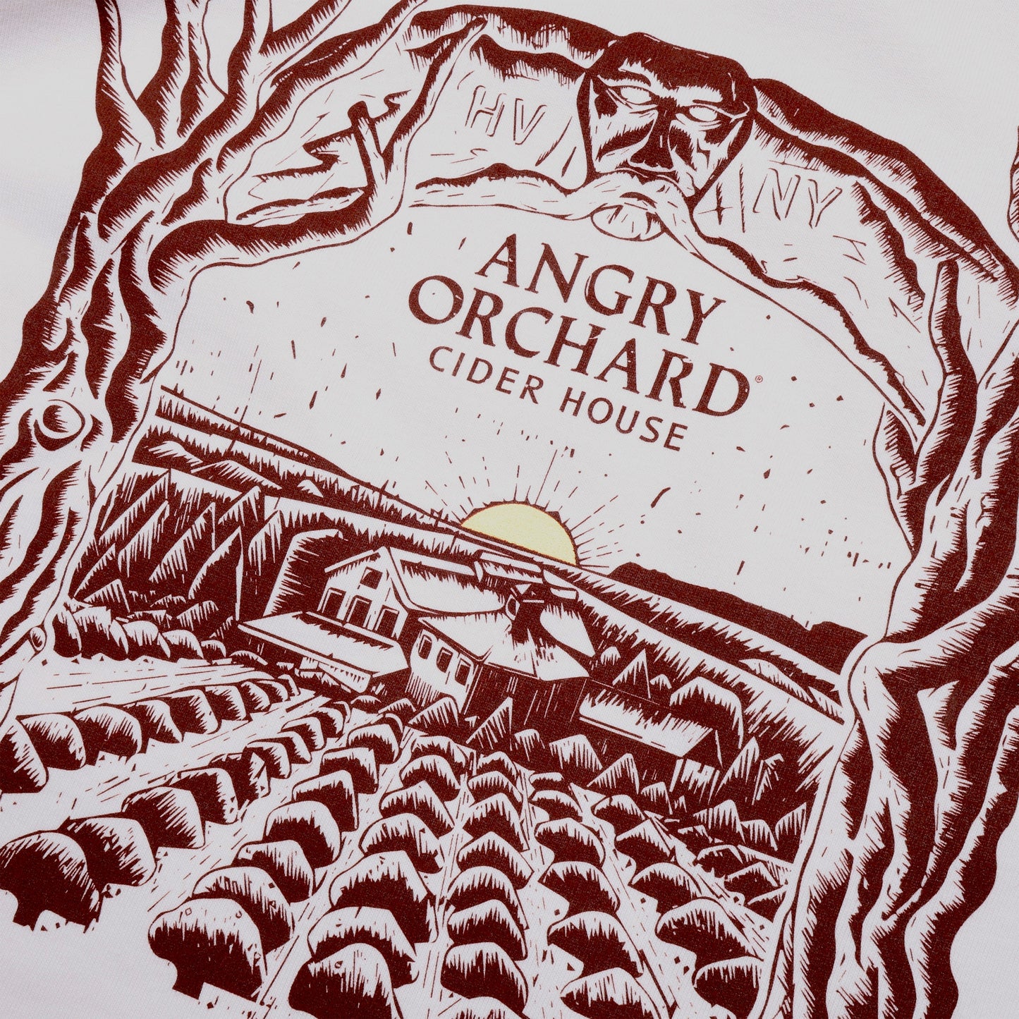 Close up of the Angry Orchard Cider House design in dark red print on a white bone short sleeve T-shirt. The design is a hand drawn like image of the cider house sitting in the orchard, with a small yellow sun setting on the horizon. Bordering the sides of this image are two trees that form an arch over the top border imagery. In the center of this imagery reads "Angry Orchard, Cider House" in the classic angry orchard font.