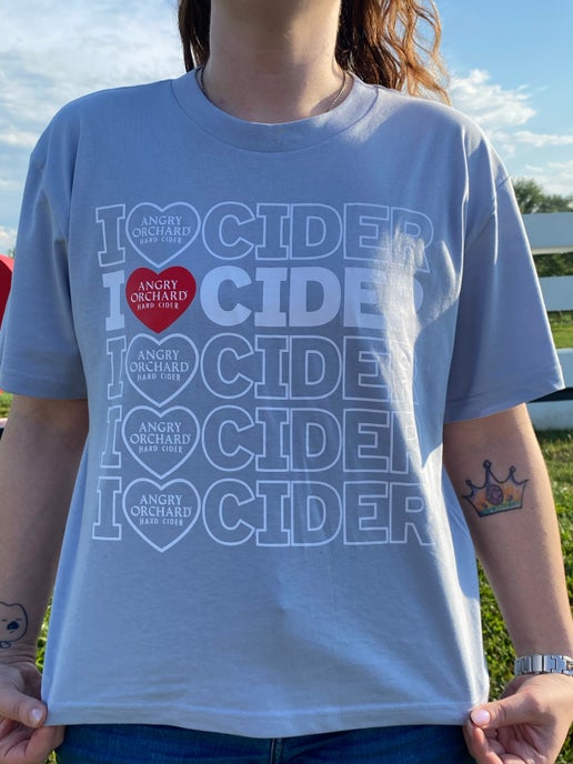 Front of gray tee displays five rows of writing "I Heart Cider". Heart is an actual heart and inside it is writing "Angry Orchard Hard Cider". Second row of writing is bolded with a red heart. 