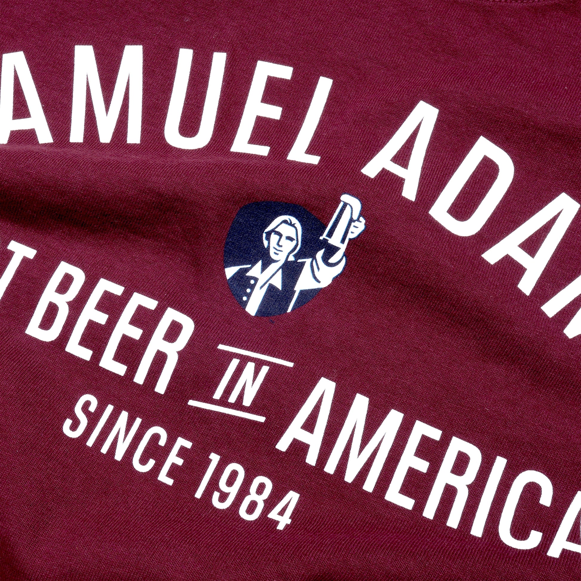 Close-up of graphics on the front. Front view of tee features writing "Samuel Adams" a small image of blue shield with Sam Adams hoisting a beer and writing "Best Beer in America Since 1984" underneath.
