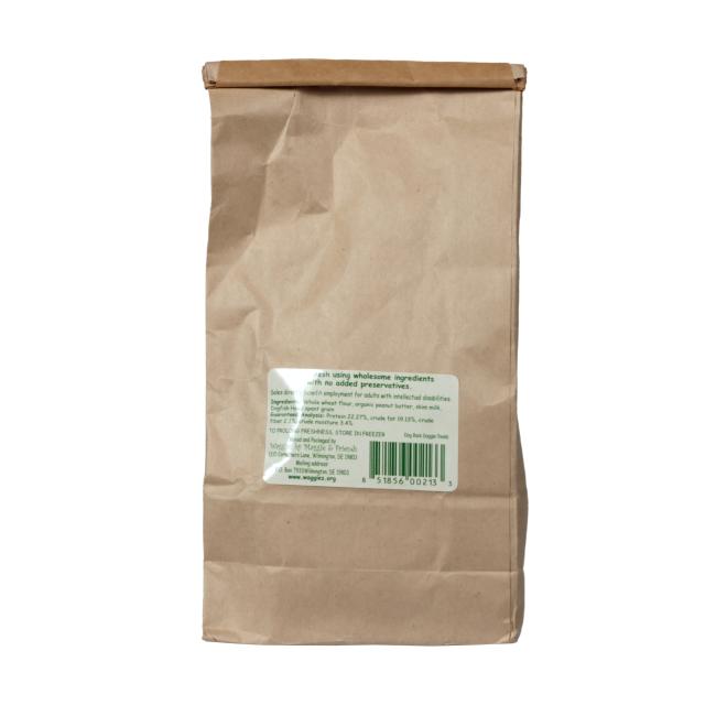 A plain brown paper bag stands upright with a folded top and a white label featuring green and black text and a barcode, possibly containing Waggies Peanut Butter Dog Treats by Waggies by Maggie & Friends Inc. The text is not fully readable.
