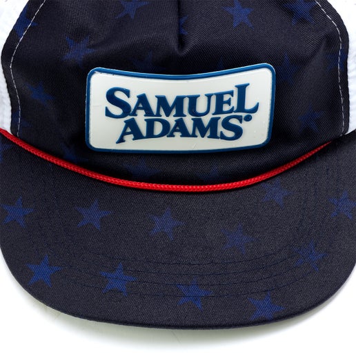 A navy trucker hat with red rope piping and a white Samuel Adams patch on the front. Reveals blue stars when wet.